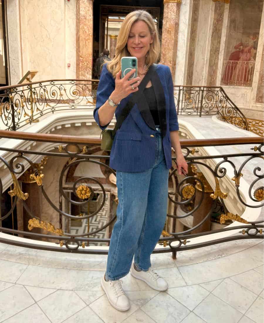 A woman stands in front of an ornate railing on a marble floor. She is wearing a blazer, jeans and white sneakers.