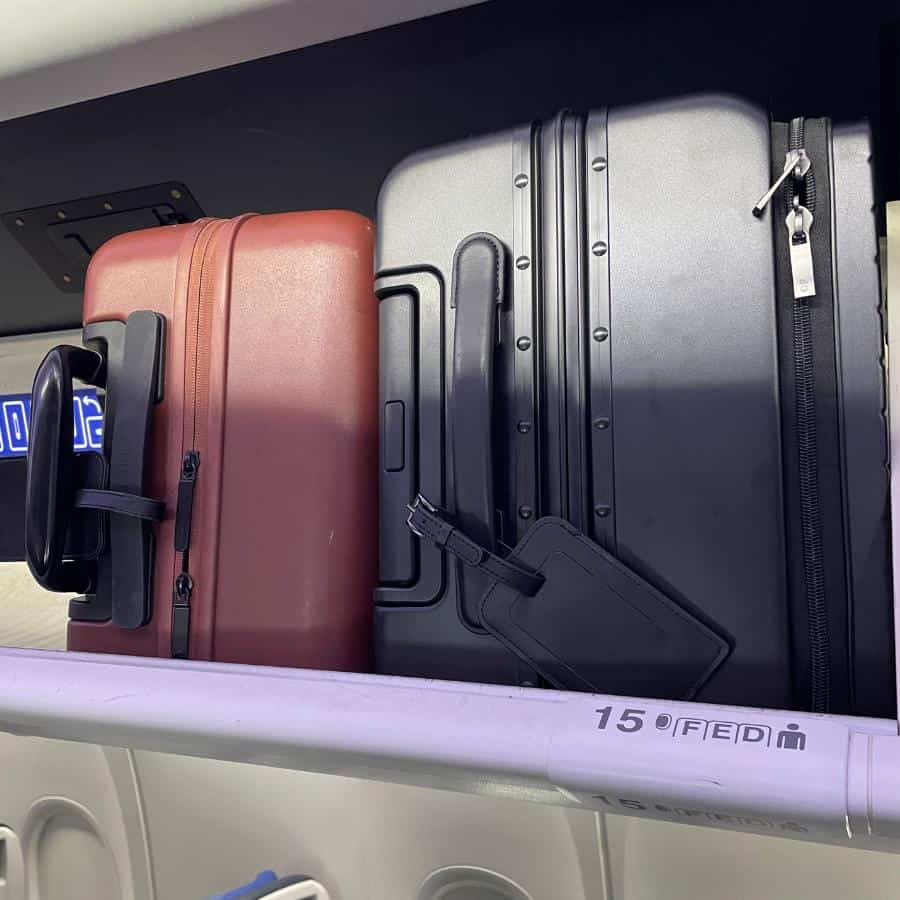 Two carry-on roller bags are visible inside an airplane overhead bin. They are placed on their narrow side — one is brown and a slightly larger one is black. The row number is visible and says 15 FED.