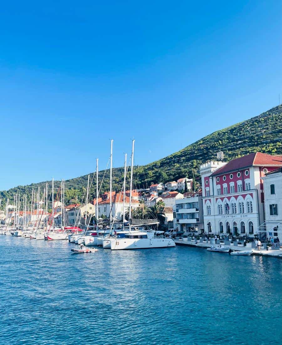 A row of sailboats are moored in the harbor of the Croatian Island of Vis. The sea is a turquoise color and the whitewashed buildings have red roofs.