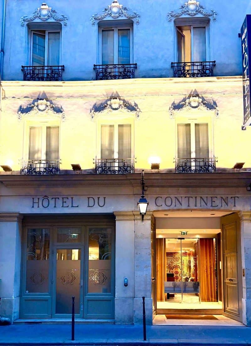 The front of a hotel in Paris shows limestone facade and traditional windows. It is evening and building is lit up.