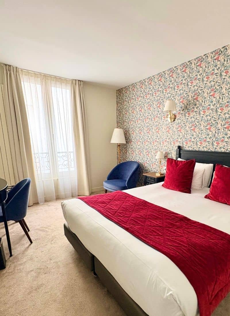 A Paris hotel room shows a double bed with white cover and red accent pillows. Traditional french windows and modern flower wallpaper with sconces. Two navy armchairs are also visible.