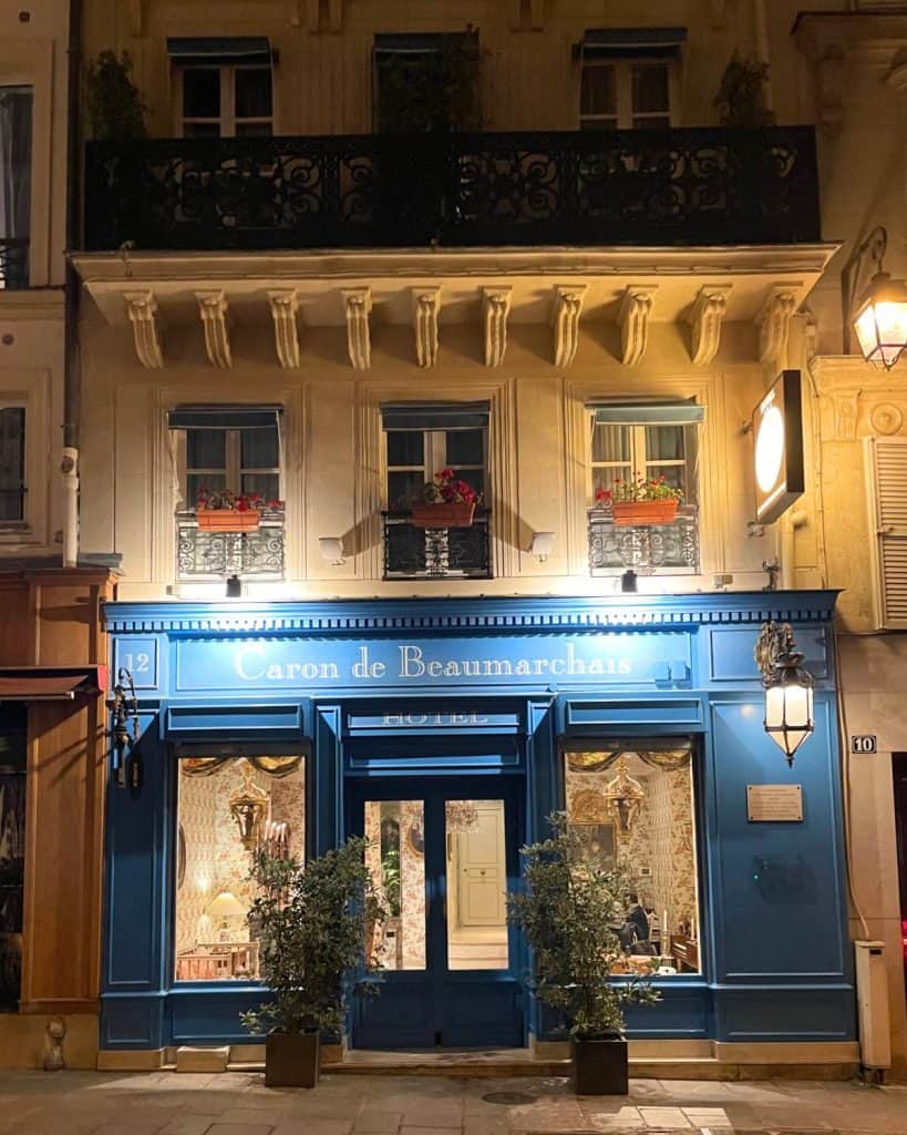 A pretty blue exterior of a hotel is set on a cobblestone street at night. A large lit lantern hangs to one side of the entry door and the lobby area inside is lit up with flowered wallpaper visible.