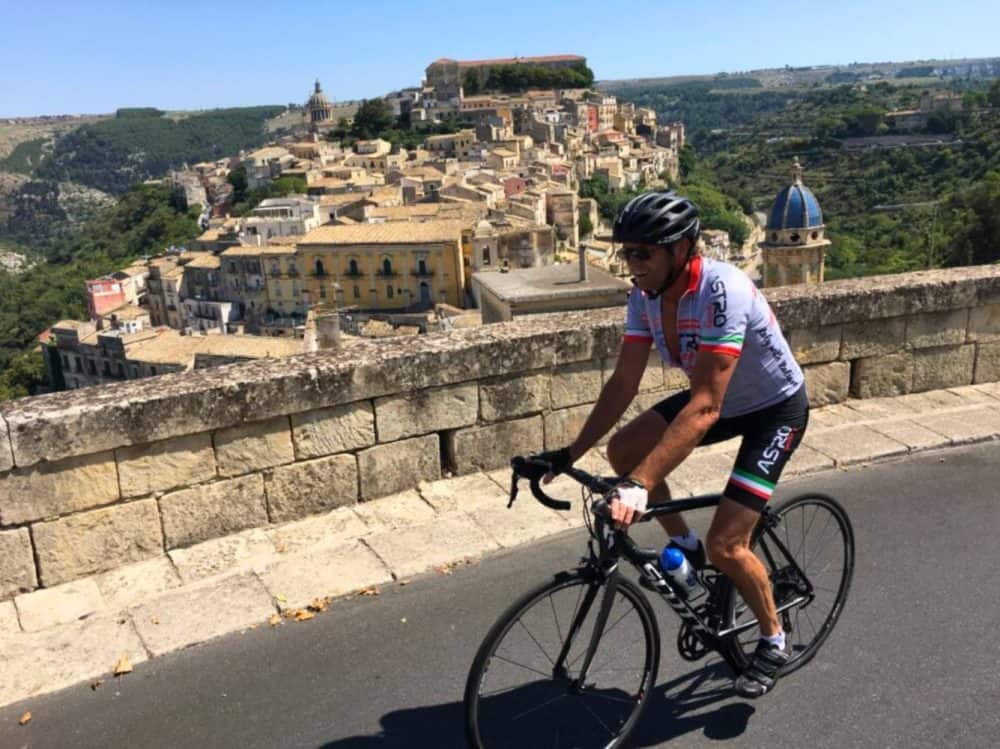 A man on a bicycle climbs a paved road with an Italian hill town visible beyond him