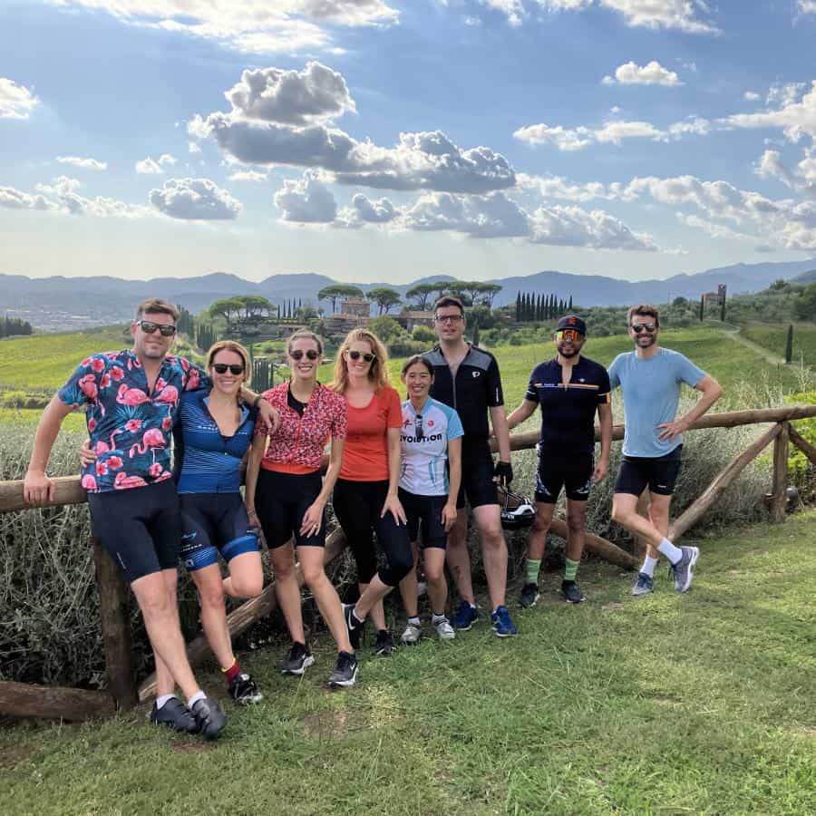 A group of cyclists stand together after a day of riding. Rolling hills and a a stone fence are behind them.