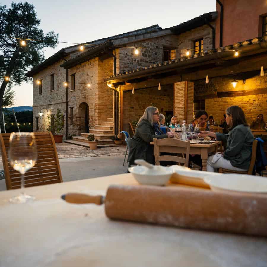 Outdoor dining in Italy next to a traditional villa. A wine glass and rolling pin sit atop a table with guests at another table just beyond.